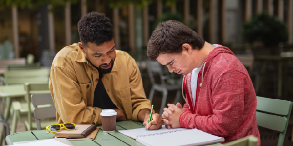 Two men sit at a café table outdoors with notebooks spread out in front of them. One man is writing in the other man’s notebook.