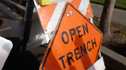 An orange road sign reads “Open Trench.” Signs of roadwork are visible behind it. 