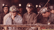 A sepia-toned color photograph showing a group of miners in the 1970s as they enter a lift that will take them underground. They are wearing helmets with headlamps. Photo credit: National Archives #556336