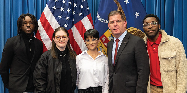 Secretary Walsh poses with four apprentices in front of U.S. and Labor Department flags.