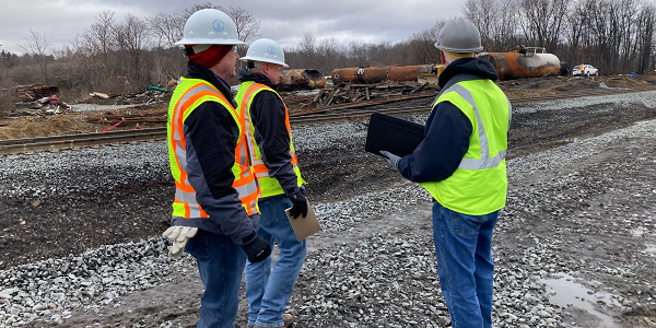 Three men in hard hats and safety vests stand near a derailed train in East Palestine, Ohio. One is taking notes. 
