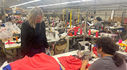 In a crowded work room, a woman pushes red fabric through a sewing machine.