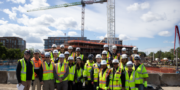 A group of people in hardhats and safety vests pose for a photo in front of two cranes at a construction site.