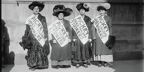 Black-and-white photo of four women garment workers striking during the "Uprising of the 20,000" in New York City, February 1910. The women are wearing sashes reading "Picket Ladies Tailors Strikers."