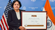 Lalitha Natarajan, wearing a dark suit, stands in front of U.S. and India flags holding a framed award.