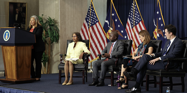 A woman speaks at a podium while a panel of four professionals sits on the stage.