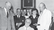 Black and white photo. President Franklin Roosevelt signs the Social Security Act, flanked by supporters, including Secretary of Labor Frances Perkins.