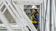 A woman in safety gear stands on a scaffold, looking out over a building site.