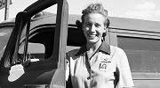 Black and white 1940s photo of a worker standing next to her truck, smiling.