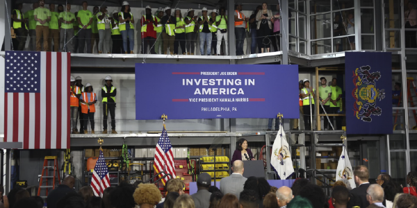 In a factory, surrounded by construction workers, Julie Su speaks at a podium under a banner reading "President Joe Biden, Vice President Kamala Harris, Investing in America"