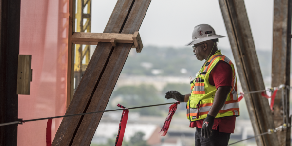 A construction worker stands on an elevated platform, near scaffolding and a line of supportive girders, gazing toward the horizon. 