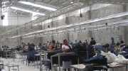 A maquiladora garment factory in Mexico. Women stand at tables in a warehouse, operating sewing machines.