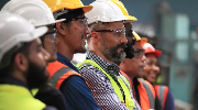 A collection of workers in safety gear stand in a line, smiling at something off camera.