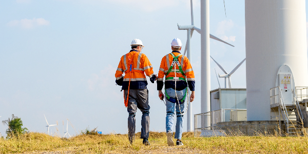 Two workers wearing high-visibility clothing, hard hats and fall protection walk near wind turbines in a field.