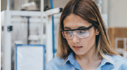 A young woman in protective goggles reads from a clipboard in a lab environment.