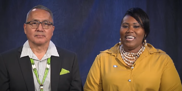 A Navajo man in a suit and a Black woman in a yellow blouse sit side-by-side, facing the camera.