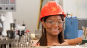 A young woman in hardhat and safety glasses smiles in a workshop.