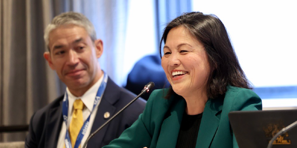 Acting Secretary Su smiles while speaking into a microphone at a roundtable. Another participant sits beside her, listening.