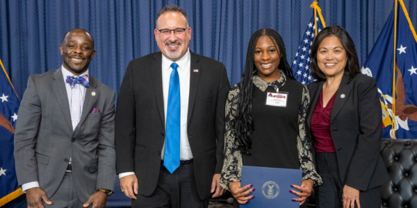 Four people, two men and two women, wearing professional business attire standing together and smiling for a photo in front of a blue curtain and the American flag. One woman holds a graduation certificate.