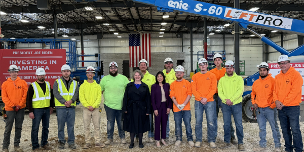 Acting Secretary Su poses for a group photo with a dozen men wearing hard hats and safety vests in a warehouse in front of an American flag.
