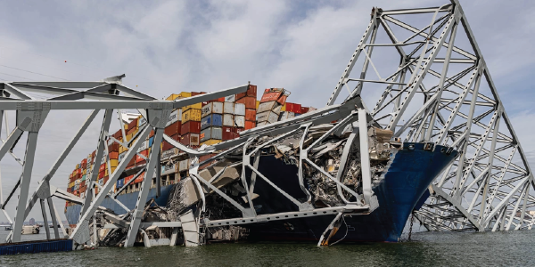 A large boat heaped with shipping containers is ensnared in the wreckage of the Key Bridge. Twisted metal wraps around its hull and rises toward the sky from the waters of the Chesapeake Bay. Photo by David Adams, U.S. Army Corps of Engineers