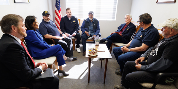 Acting Secretary Su and MSHA Assistant Secretary Chris Williamson sit in a small room with several miners and union officials. An American flag is behind them, and light is streaming in from a large window.