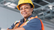 A Black woman wearing protective gear and a hard hat smiles with her arms folded across her chest.