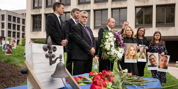 Assistant Secretaries Doug Parker and Chris Williamson stand outside with family members of workers killed on the job, all in dark clothing. Several hold large photos of lost loved ones, standing by a wreath and table with individual roses and a bell.