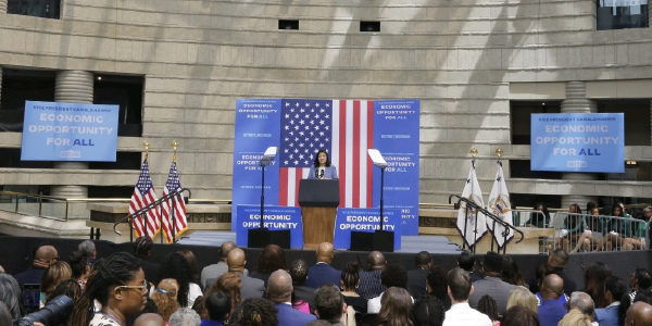 Acting Secretary Su speaks from a stage in front of American flags and signs reading “Economic opportunity for all.”