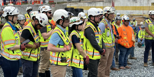 Construction workers in safety vests and hardhats stand at a construction site, listening to a speaker.