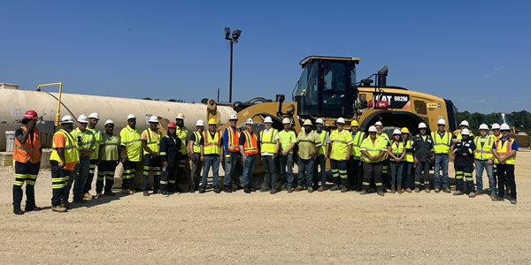 MSHA staff stand outside on a cloudless day at a mine in Maryland with about two dozen mine workers. All wear high visibility clothing and hardhats. A large bulldozer and cylindrical concrete container are behind them. 