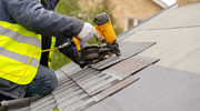 Close-up of a roofing worker wearing a reflective jacket and gloves using a nail gun to install shingles.