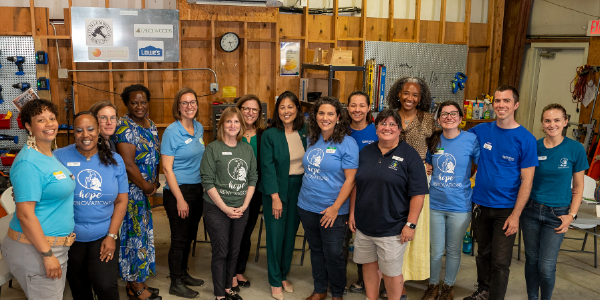 Acting Secretary Su poses with a dozen women, many wearing Hope Renovations tee shirts. Ladders and drills are visible in the workshop behind them. 