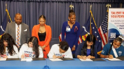 Five young people sign documents at a table while three adults look on.