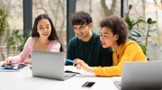 Three young people working with a laptop and notebooks at a table, smiling and engaged in their task. The setting appears to be a bright room with large windows. 