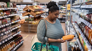 A woman holding a shopping basket looks at a bottle of juice in a grocery store refrigerator aisle. 