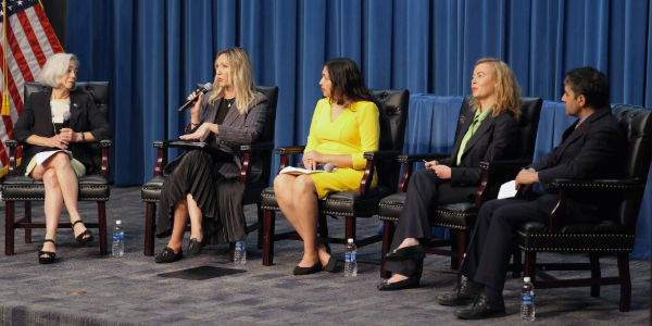Thea Lee moderates a panel with four other participants. They all sit in chairs on a stage, holding microphones. 