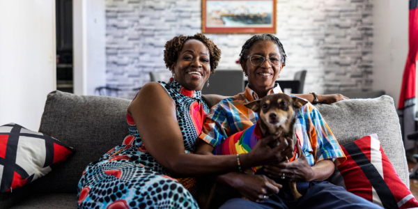 Two people sit on a couch holding a dog wearing a rainbow onesie.  