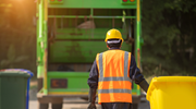 A worker in an orange high visibility vest and hard hat pulls a bin toward a green garbage truck.