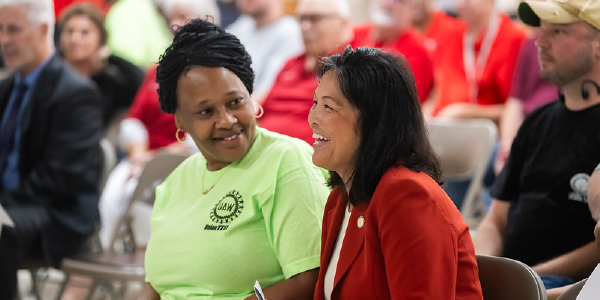 Seated in an audience on a folding chair, Acting Secretary Su laughs. Sitting next to her, a Black woman in a UAW shirt looks at Su with a smile. 