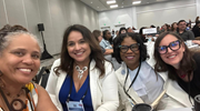 Four diverse women in business clothes smile for a selfie in a conference room.