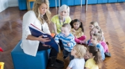 A woman shows a book to a group of toddlers while another woman looks on in a school or daycare setting. 