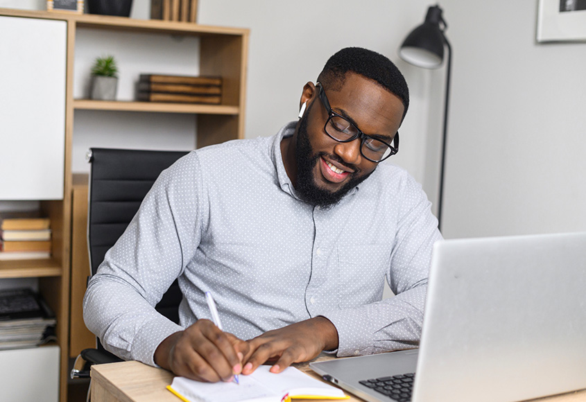 A black man writing in a notebook beside his laptop on the desk.