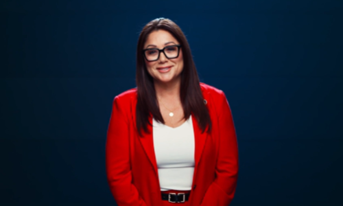 Secretary Chavez-DeRemer, wearing a red suit, stands smiling against a dark blue background