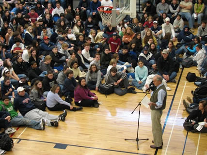 Image of dozens of students at a school assembly listening to a speaker at a microphone